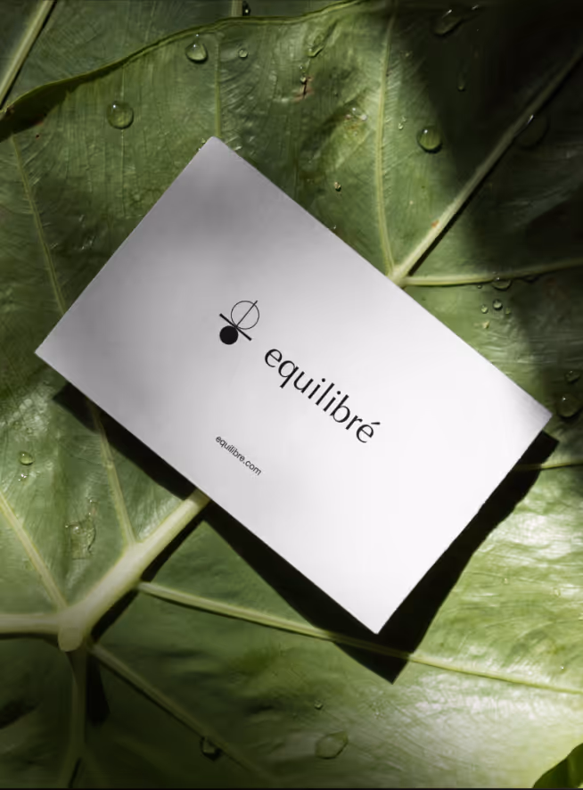 White business card with the text 'equilibré' and a simple logo placed on large green leaves with water droplets.